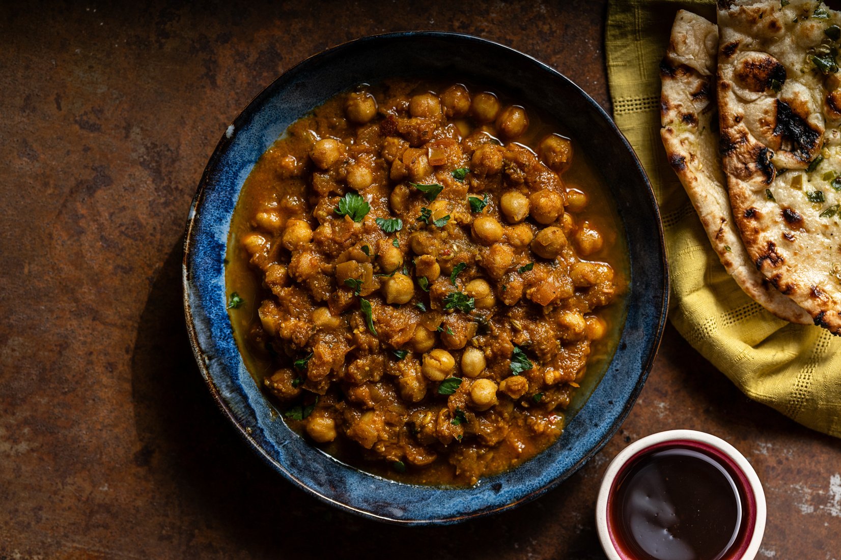 chana masala garnished with fresh parsley in a blue bowl, with garlic naan on a yellow napkin on the right side